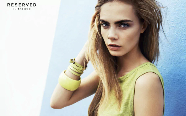 HD desktop wallpaper featuring English actress and model Cara Delevingne with blue eyes and blonde hair against a blue background, styled in a green sleeveless top.