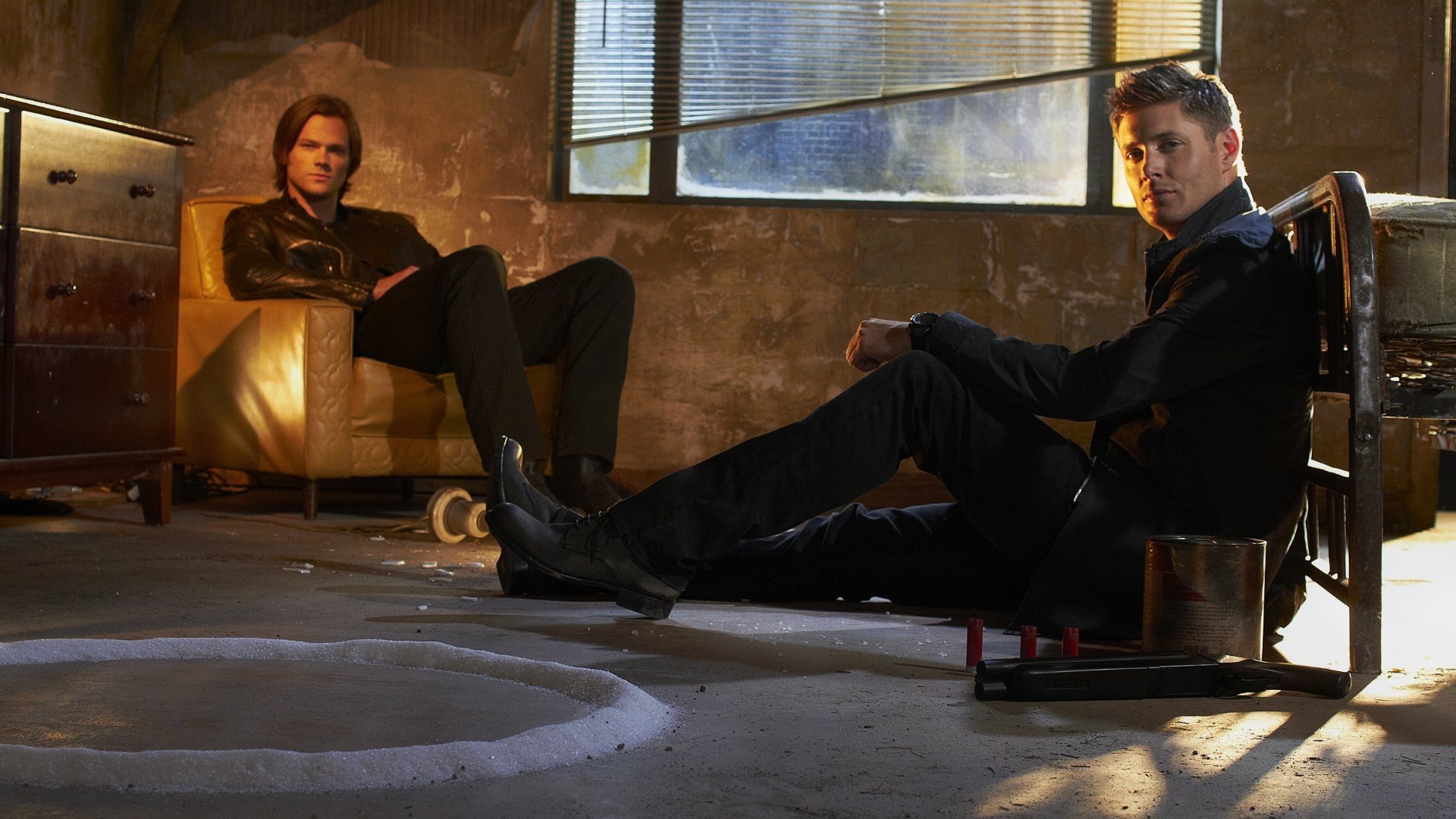 A dramatic scene from the TV show Supernatural, featuring two main characters seated in a dimly lit room, with a mysterious circle on the floor, creating a tense atmosphere.