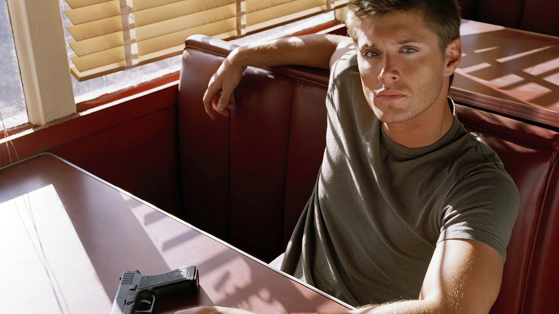 A character from the TV show Supernatural rests casually in a diner booth, with a relaxed pose and a handgun beside him, capturing a moment of intensity and intrigue.