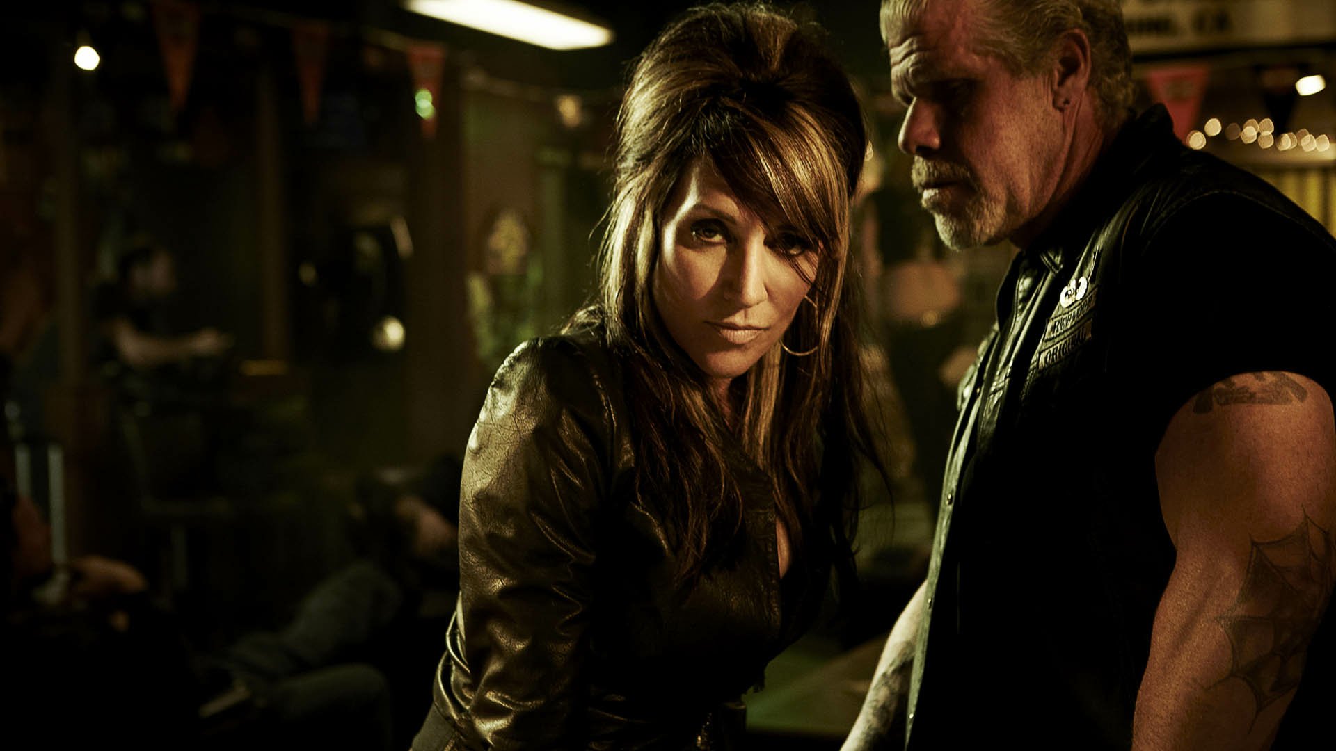 HD PC desktop wallpaper for the TV show Sons of Anarchy: two leather-clad club members in a dim, smoky bar — a woman staring intently at the viewer with a man standing beside her.