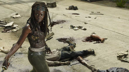 Michonne from *The Walking Dead*, portrayed by Danai Gurira, stands confidently amidst a post-apocalyptic scene littered with fallen foes. HD wallpaper highlighting her fierce character.