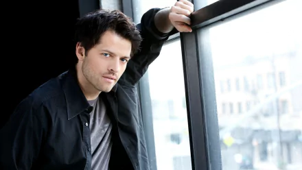 A striking HD wallpaper featuring a character from the TV show Supernatural, gazing thoughtfully while leaning against a large window with an urban backdrop.