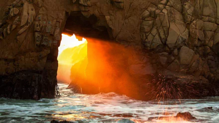 A stunning sunbeam streams through a rocky arch along the coastline, casting a warm glow over the waves and highlighting the beauty of nature in this HD wallpaper.
