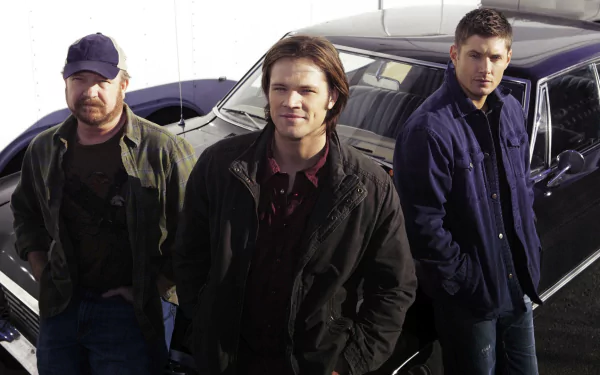 Three main characters from the TV show Supernatural pose confidently in front of a classic car, showcasing a blend of mystery and adventure in this HD desktop wallpaper.