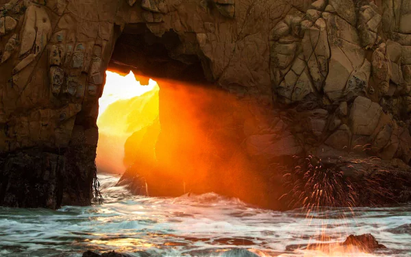 A stunning sunbeam streams through a rocky arch along the coastline, casting a warm glow over the waves and highlighting the beauty of nature in this HD wallpaper.