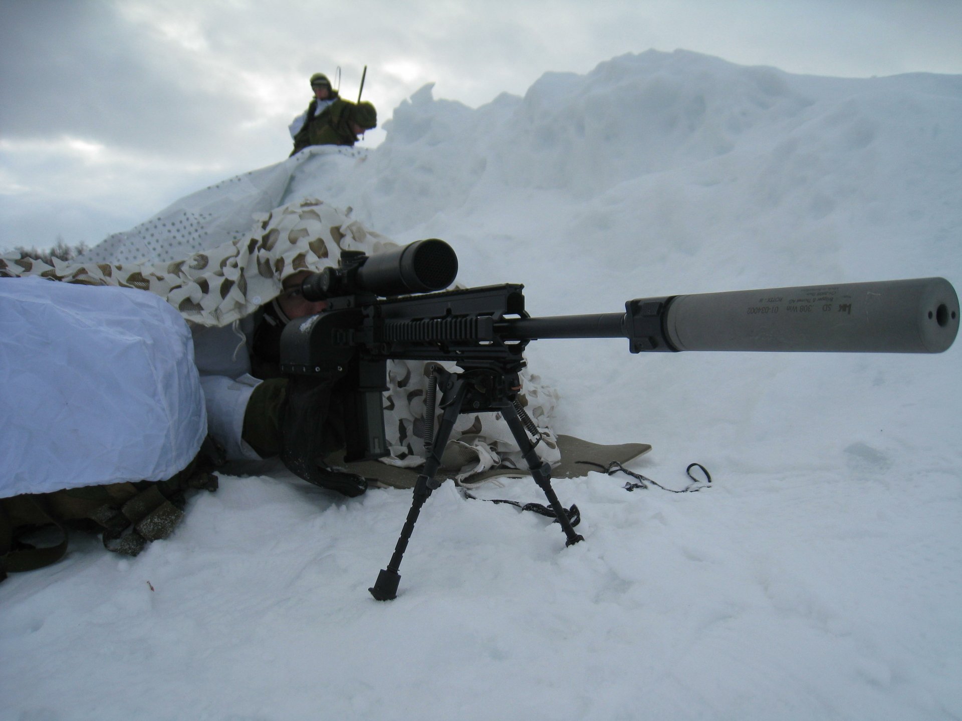 HD desktop wallpaper showing a military sniper positioned in snowy terrain, aiming through a camouflaged rifle with a suppressor attached.