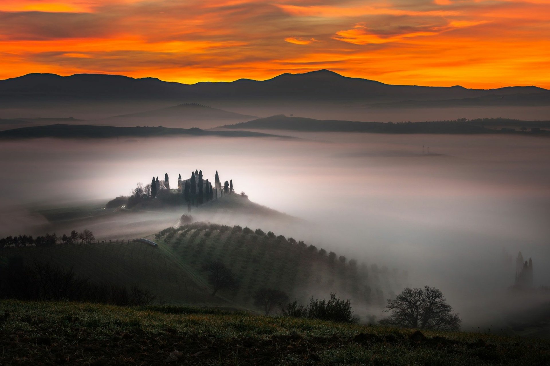 HD landscape photography wallpaper of foggy Tuscany, Italy — cypress-topped hill and vineyard rows emerging from morning mist beneath a glowing orange sky.