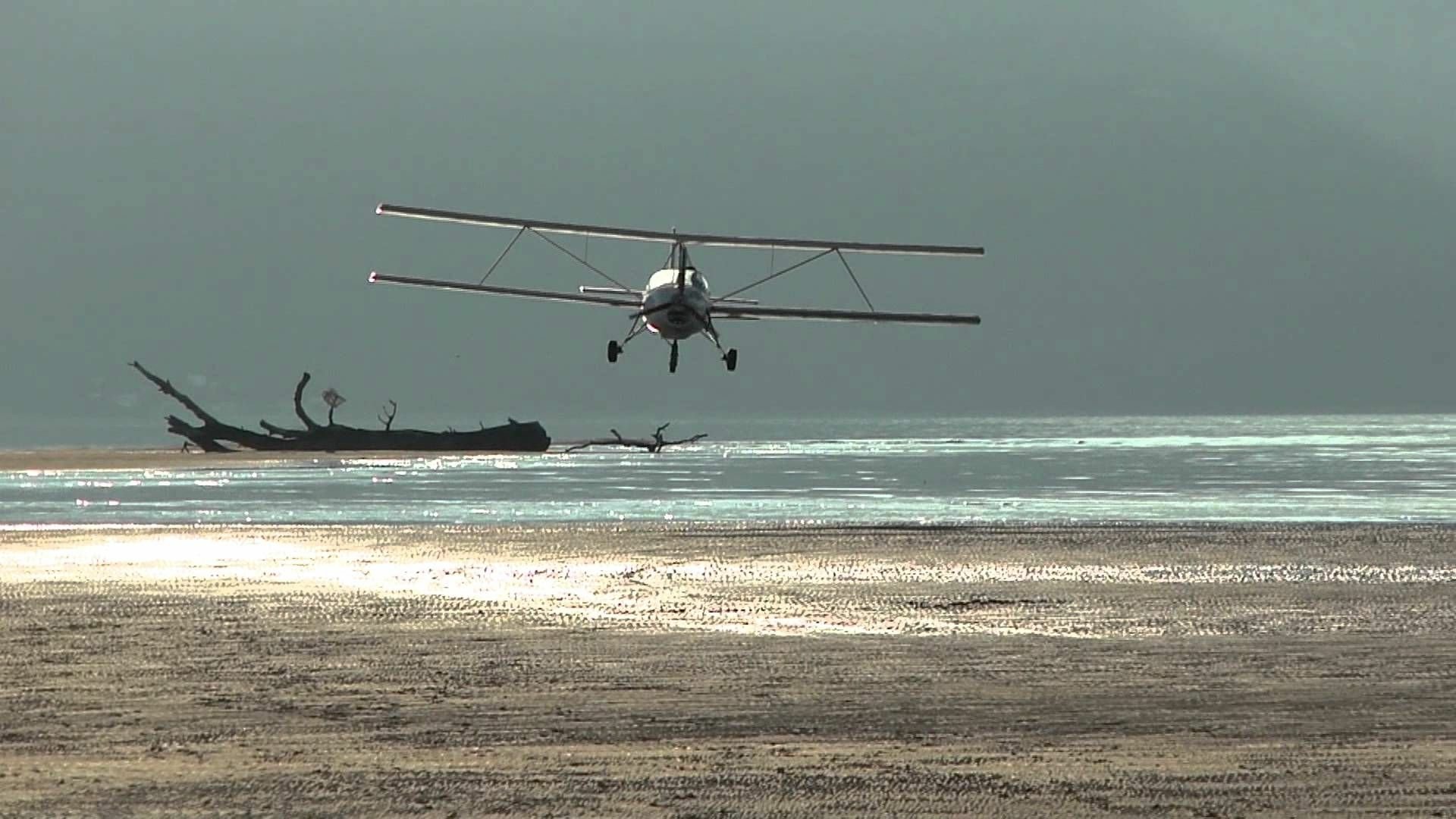 Aviasud Mistral biplane flying low over a sunlit beach and shallow sea, presented as an HD PC desktop wallpaper/background.