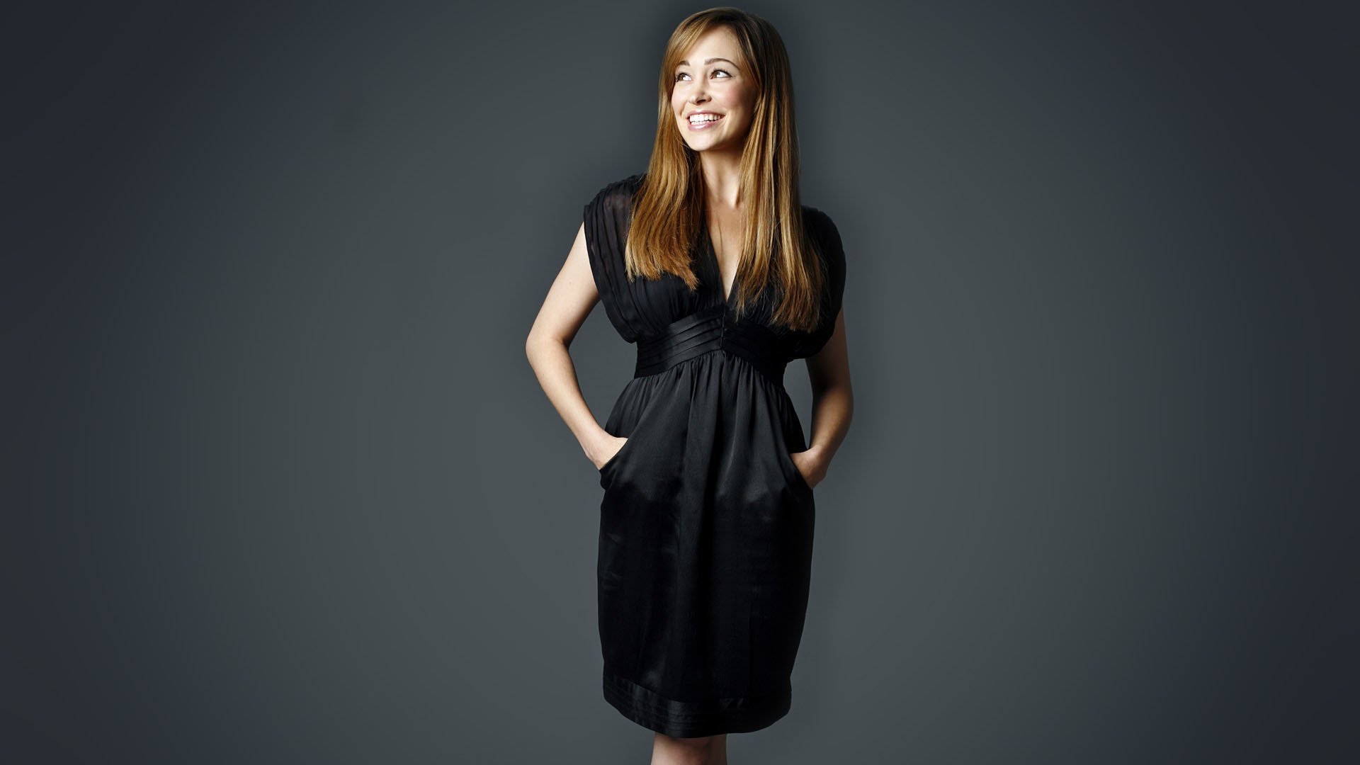 HD PC desktop wallpaper of a smiling female celebrity in a black dress, hands in pockets, standing against a smooth neutral gray studio background.