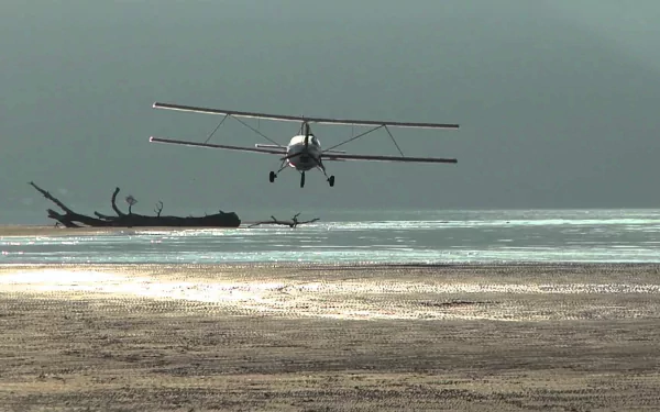 Aviasud Mistral biplane flying low over a sunlit beach and shallow sea, presented as an HD PC desktop wallpaper/background.
