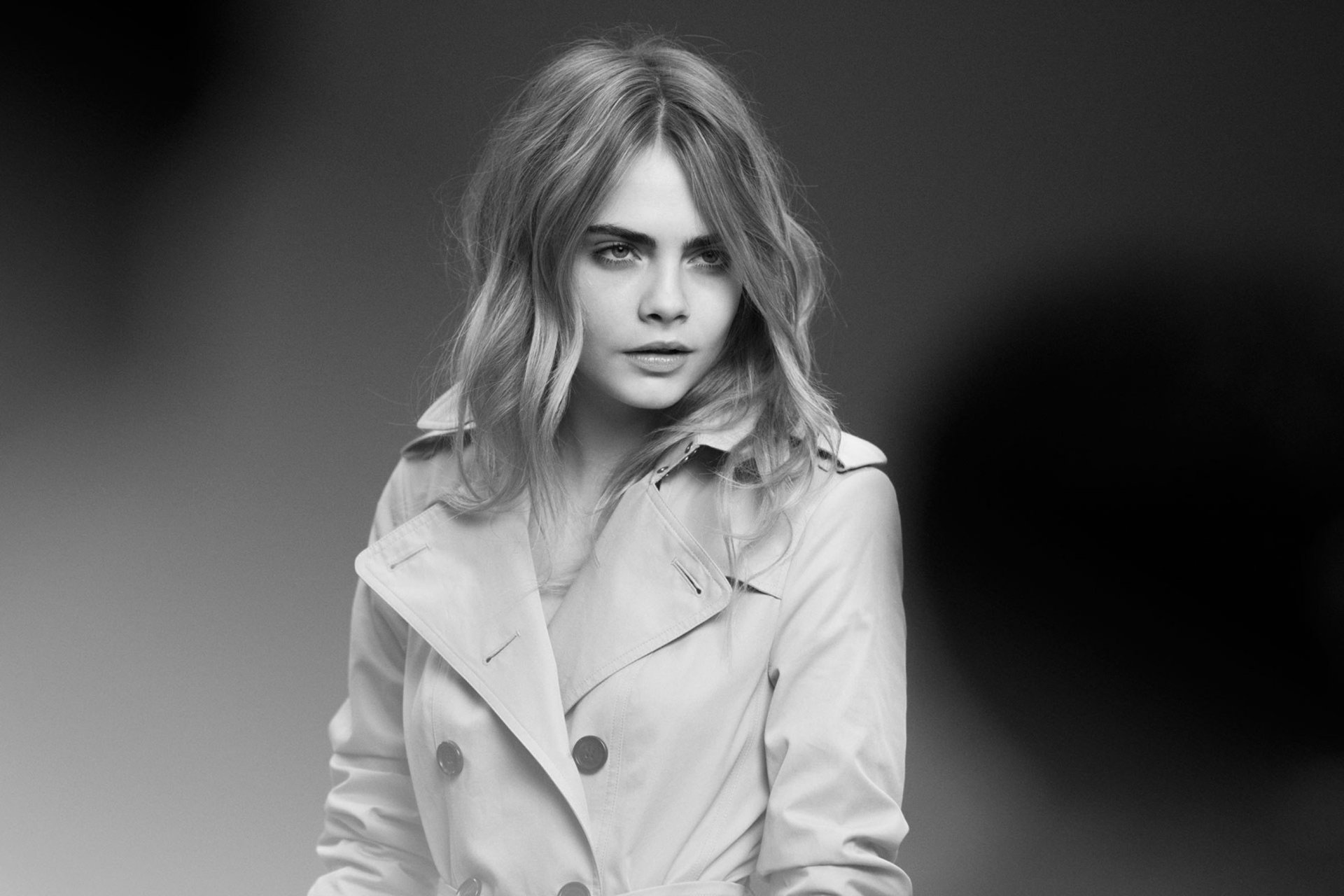 A monochrome HD desktop wallpaper featuring model and actress Cara Delevingne, showcasing her stylish look in a trench coat against a soft-focus background.