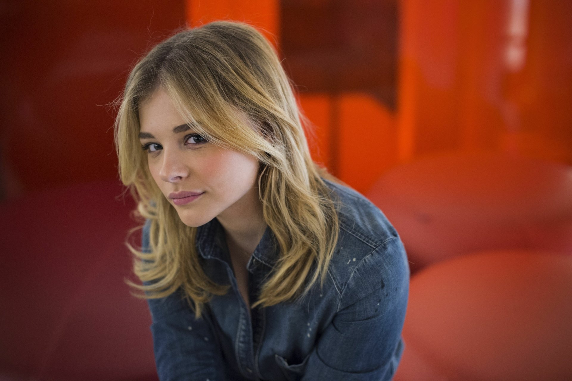 Chloë Grace Moretz poses thoughtfully against a vibrant orange background, showcasing her distinctive style in this striking 4K Ultra HD desktop wallpaper.