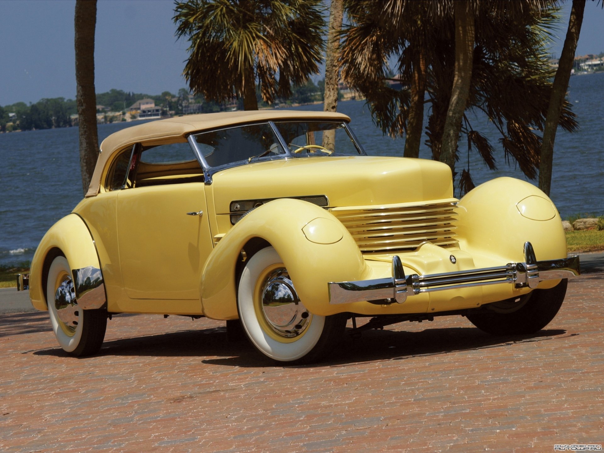 HD desktop wallpaper showcasing a classic yellow Cord 812 SC Phaeton parked by a waterfront with palm trees in the background.