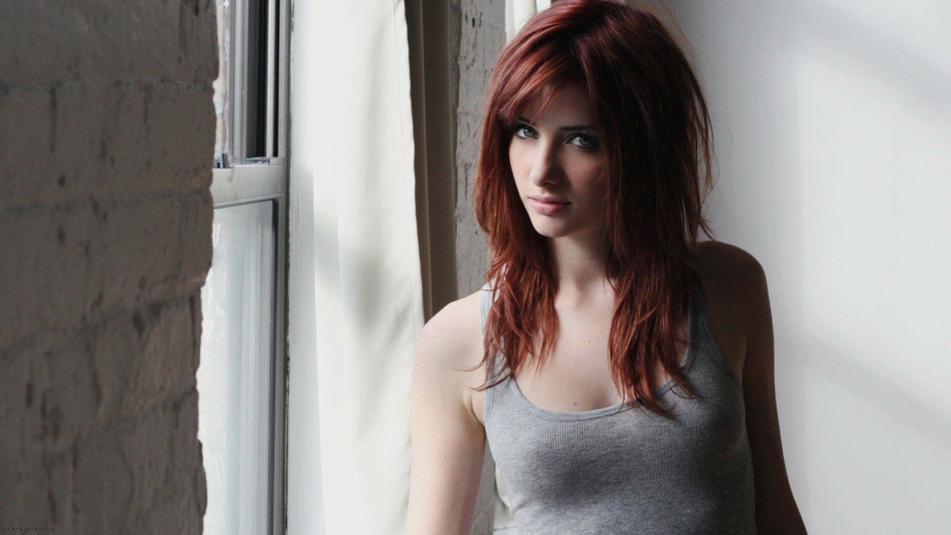 HD desktop wallpaper featuring celebrity Susan Coffey with red hair, wearing a gray tank top, standing by a window with soft natural light.