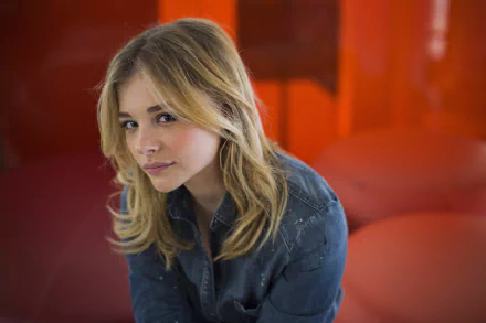 Chloë Grace Moretz poses thoughtfully against a vibrant orange background, showcasing her distinctive style in this striking 4K Ultra HD desktop wallpaper.