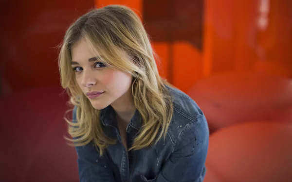 Chloë Grace Moretz poses thoughtfully against a vibrant orange background, showcasing her distinctive style in this striking 4K Ultra HD desktop wallpaper.