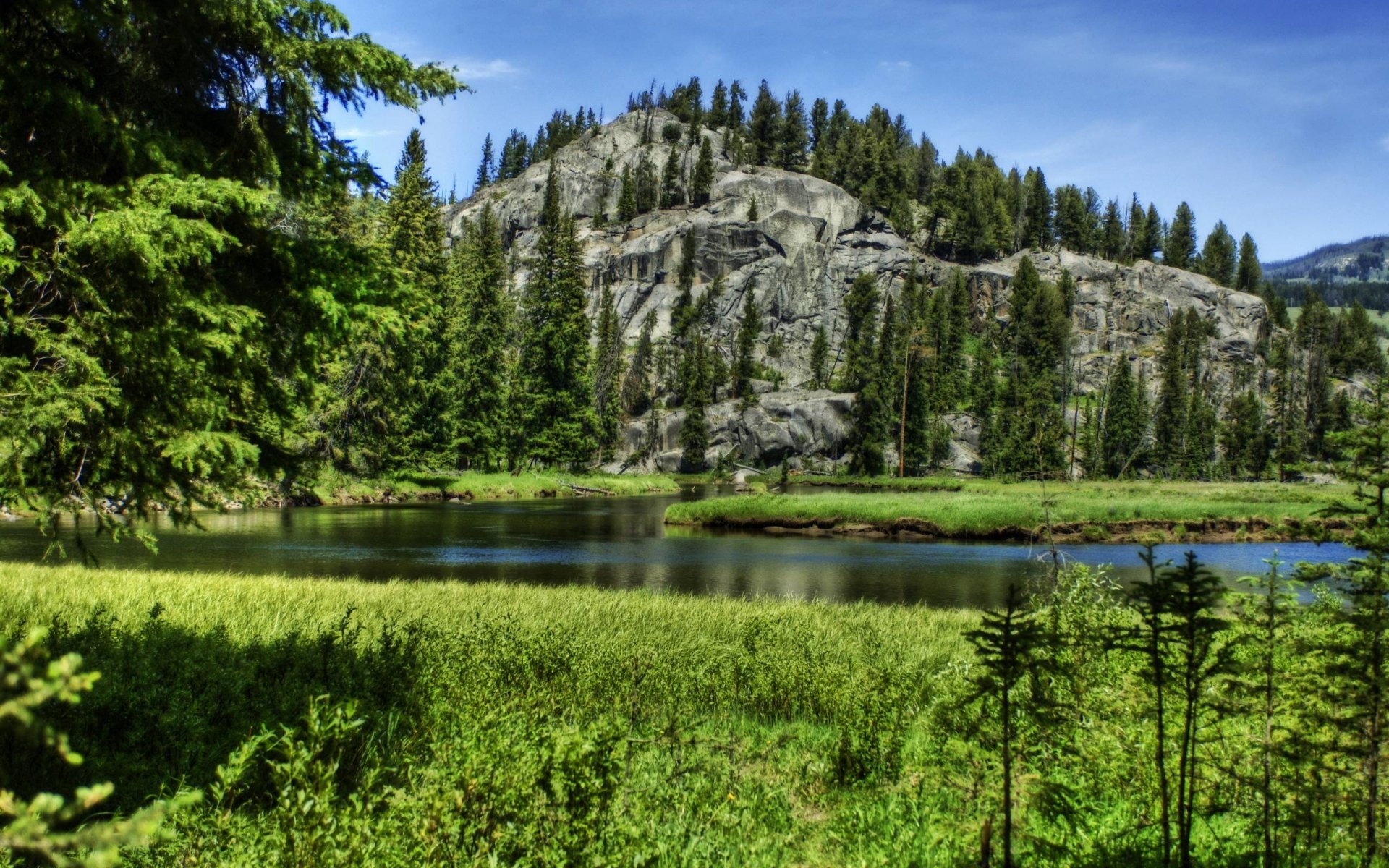 HD desktop wallpaper featuring a serene lake surrounded by lush greenery, tall trees, and a majestic rocky mountain under a clear blue sky.