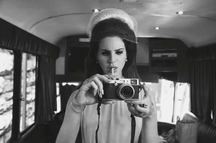 A black-and-white HD desktop wallpaper featuring Lana Del Rey, poised with a vintage camera, capturing a moment inside a stylishly decorated van.