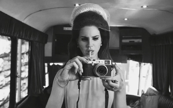 A black-and-white HD desktop wallpaper featuring Lana Del Rey, poised with a vintage camera, capturing a moment inside a stylishly decorated van.