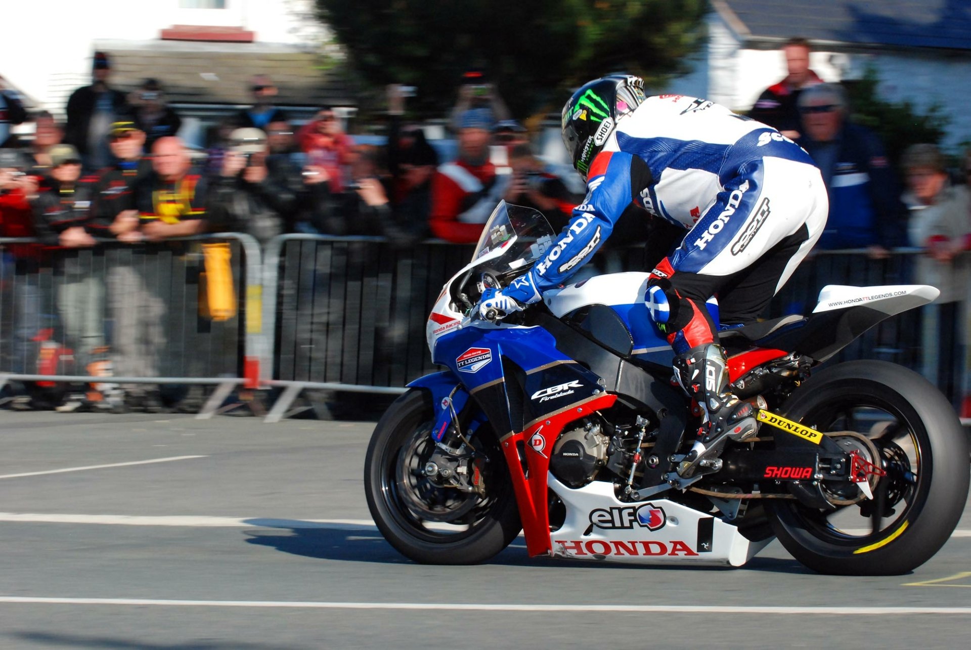 A rider in a racing suit accelerates on a Honda CBR motorcycle, surrounded by spectators at a motorsport event. The image serves as an HD desktop wallpaper.
