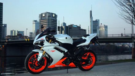 HD desktop wallpaper featuring a Yamaha YZF-R1 motorcycle parked by a riverside with a city skyline in the background during dusk.