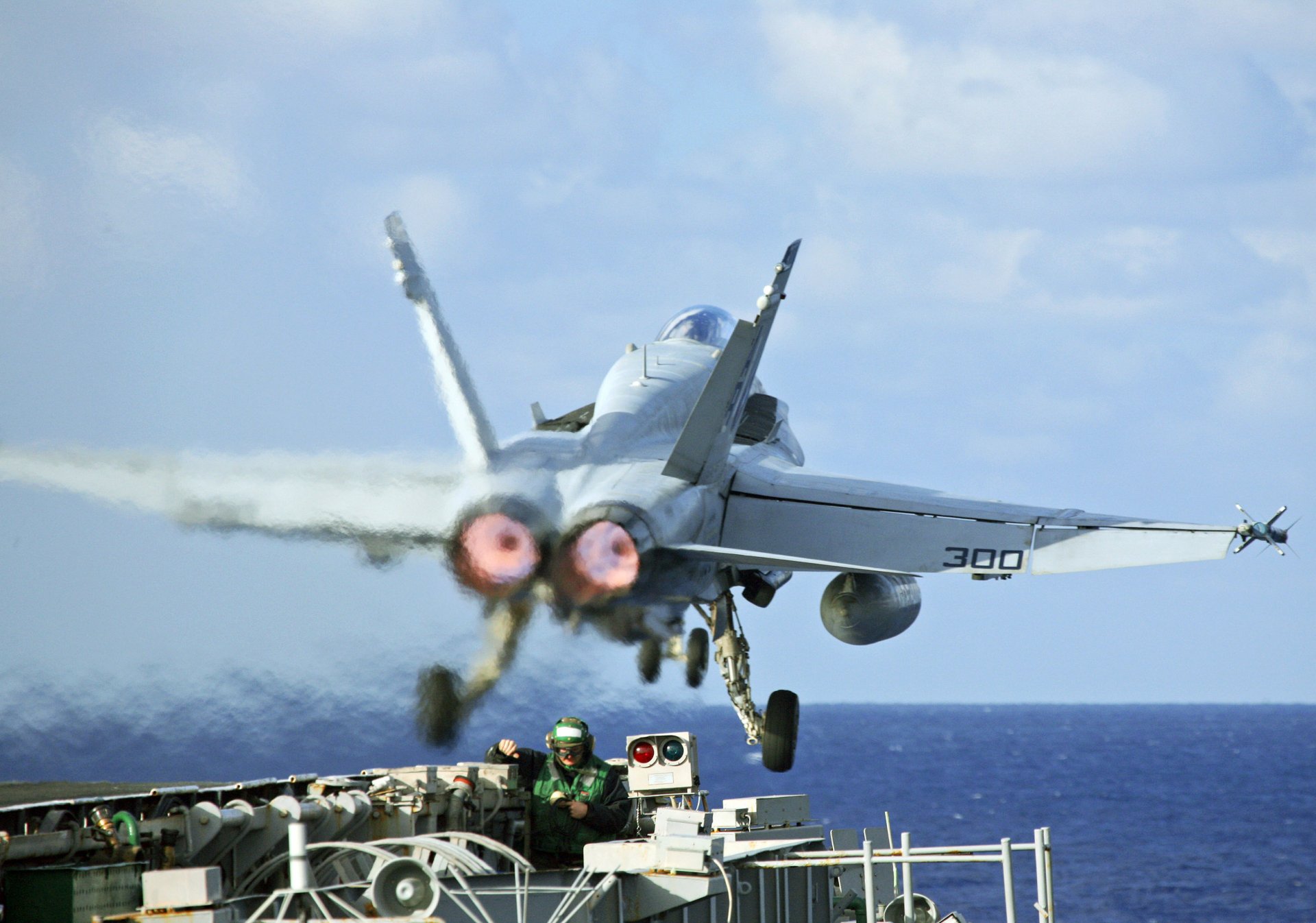 A McDonnell Douglas F/A-18 Hornet takes off from a naval ship, with military personnel in the foreground, set against a vibrant blue sky and ocean backdrop.