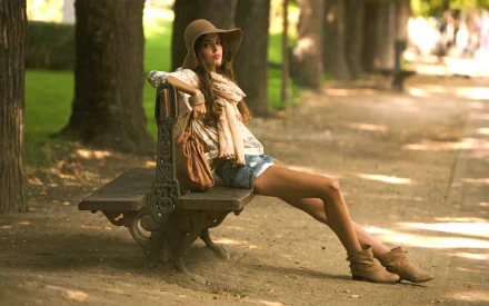 HD PC desktop wallpaper featuring a woman, Clara Alonso, sitting on a park bench wearing a hat and casual outfit in a sunlit, tree-lined pathway setting.