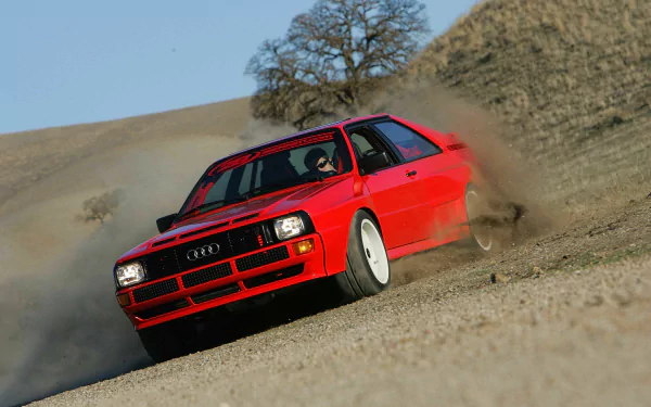 HD desktop wallpaper featuring a red Audi Quattro drifting on a dusty, hillside dirt road with a tree and dry landscape in the background.