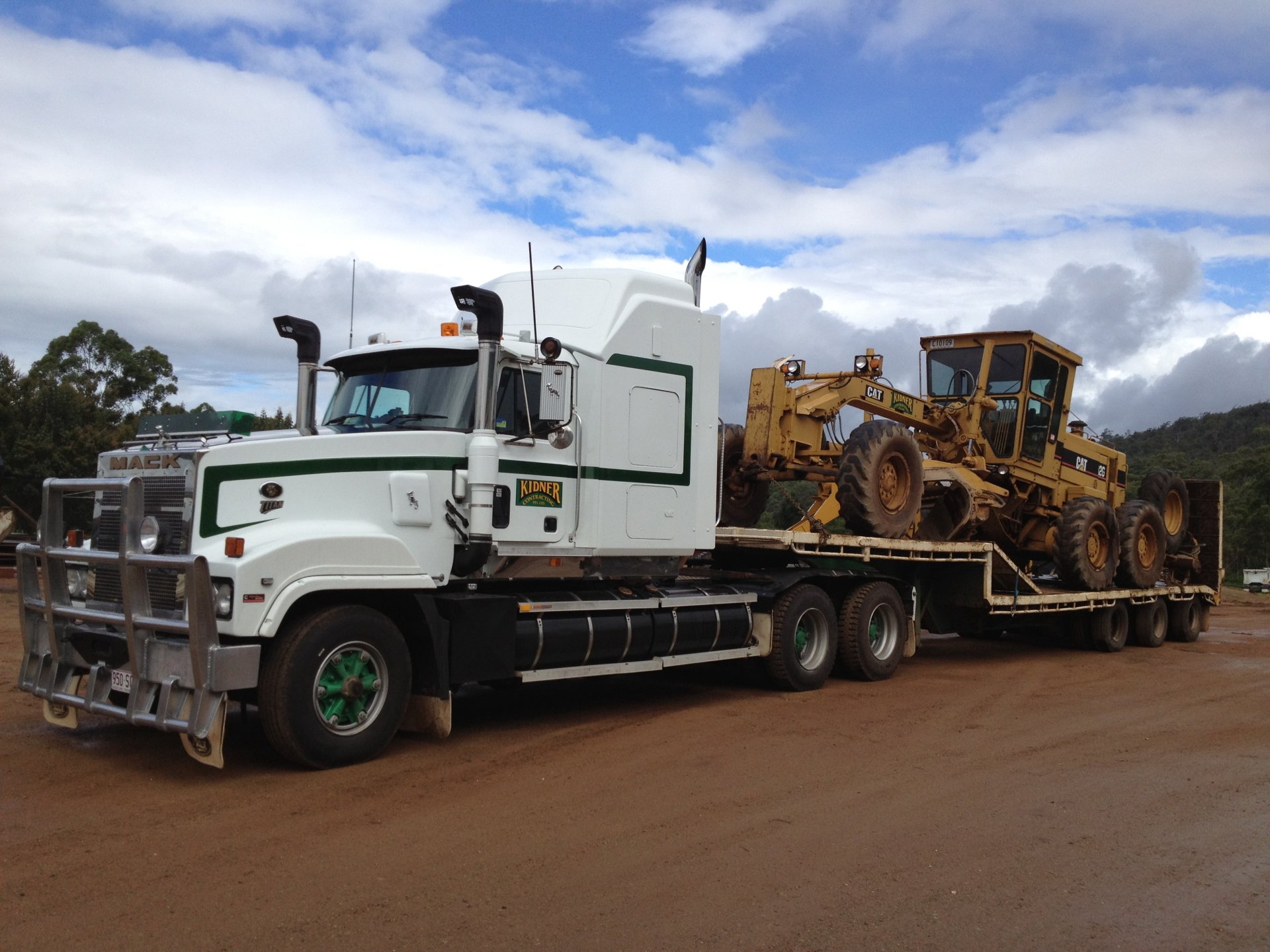 2K Quad HD PC desktop wallpaper: white Mack truck hauling a yellow grader on a flatbed trailer along a dirt road under a blue, partly cloudy sky.