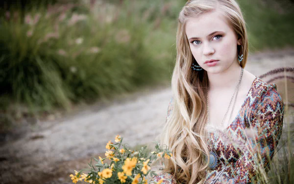 HD desktop wallpaper featuring a young woman with long blonde hair sitting outdoors, holding yellow flowers, set against a natural, blurred background.
