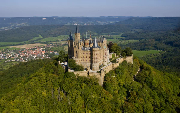 Aerial view of Hohenzollern Castle, a man-made fortress perched atop a forested hill in Germany, featured as an HD PC desktop wallpaper and background.