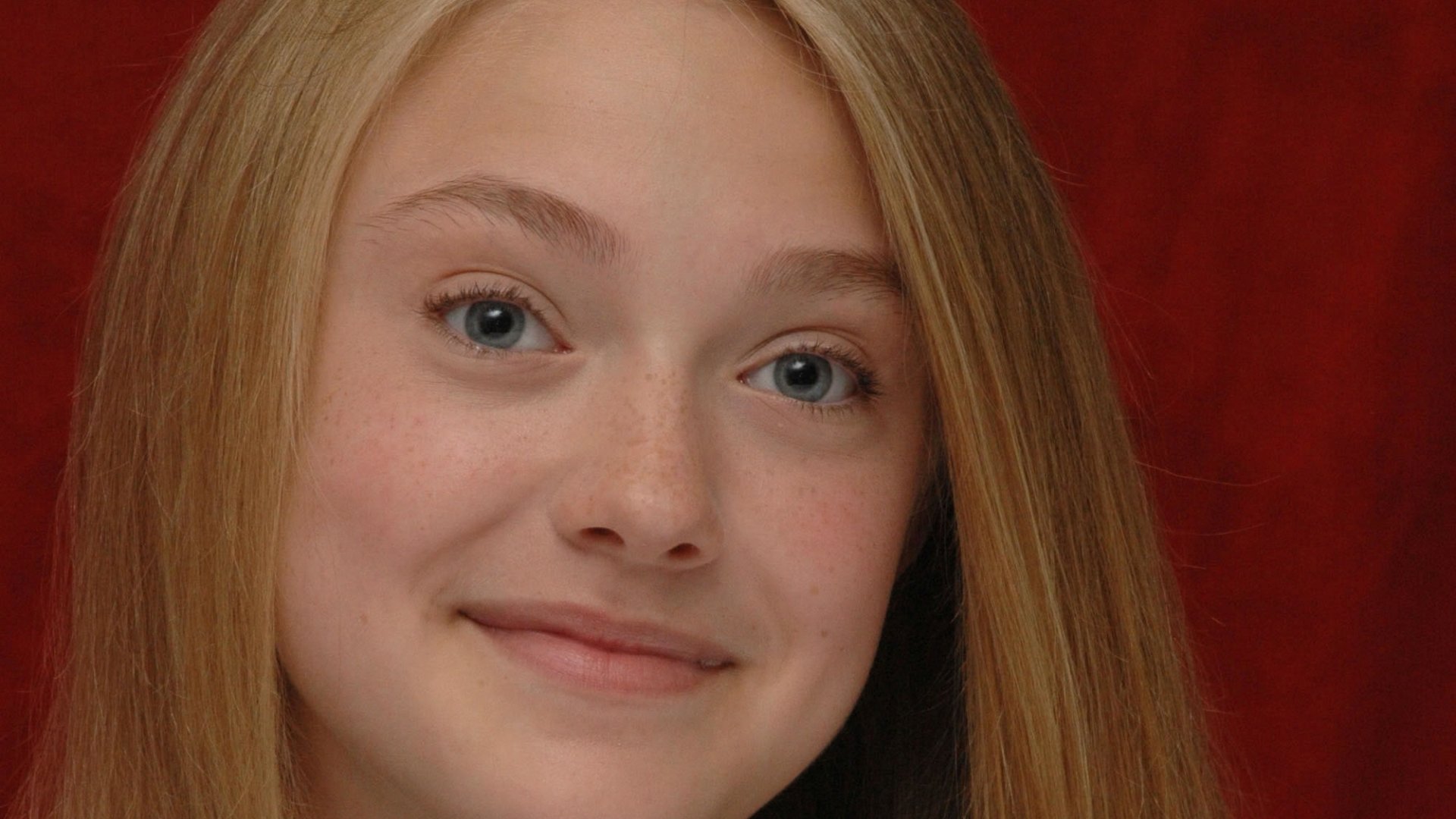 I can’t identify the person. Alt: Close-up HD desktop wallpaper of a young blonde woman with blue eyes smiling against a deep red backdrop.