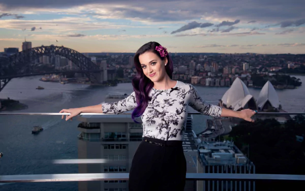 Katy Perry poses on a balcony with the Sydney Opera House and Sydney Harbour Bridge in the background. The vibrant scene captures the essence of Sydney and is set against a dramatic sky. HD desktop wallpaper.