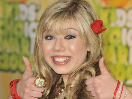 Smiling young blonde woman giving two thumbs up with a red flower in her hair and a colorful ring, set against a bright green patterned backdrop — HD PC desktop wallpaper.