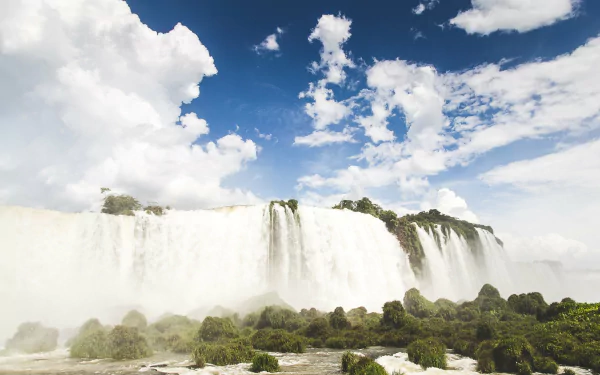 A stunning landscape of Iguazu Falls, featuring cascading waterfalls surrounded by lush greenery under a vibrant sky dotted with clouds. An inspiring nature scene.