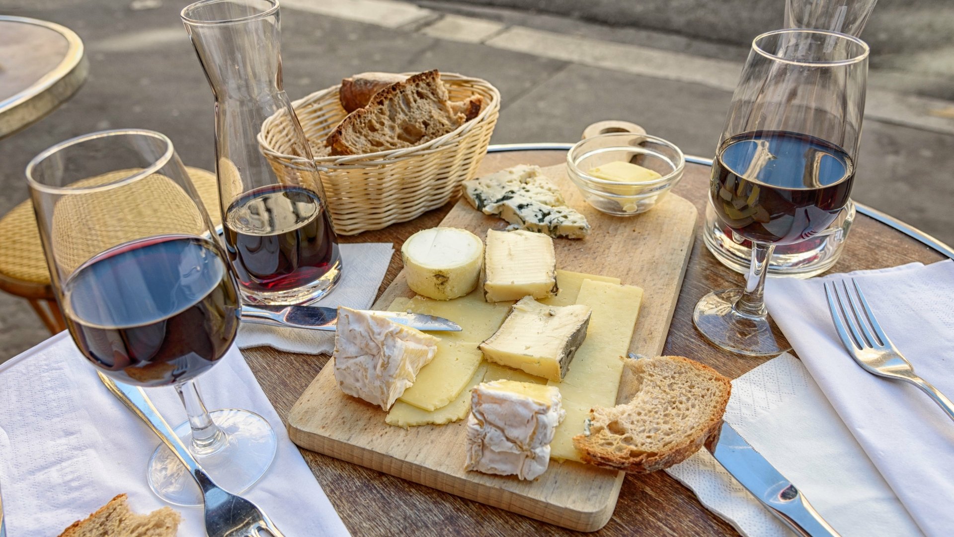A 4K Ultra HD still life of a wooden table set with glasses of red wine, assorted cheeses on a board, a basket of bread, and cutlery arranged for a meal.