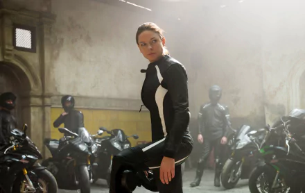 Ilsa Faust, portrayed by Rebecca Ferguson in *Mission: Impossible - Rogue Nation*, stands confidently among a lineup of motorcycles in a dramatic, industrial setting. HD wallpaper.