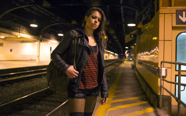 HD PC desktop wallpaper: lone woman in a leather jacket standing on a dim, empty train platform at night — moody Orphan Black TV show background.