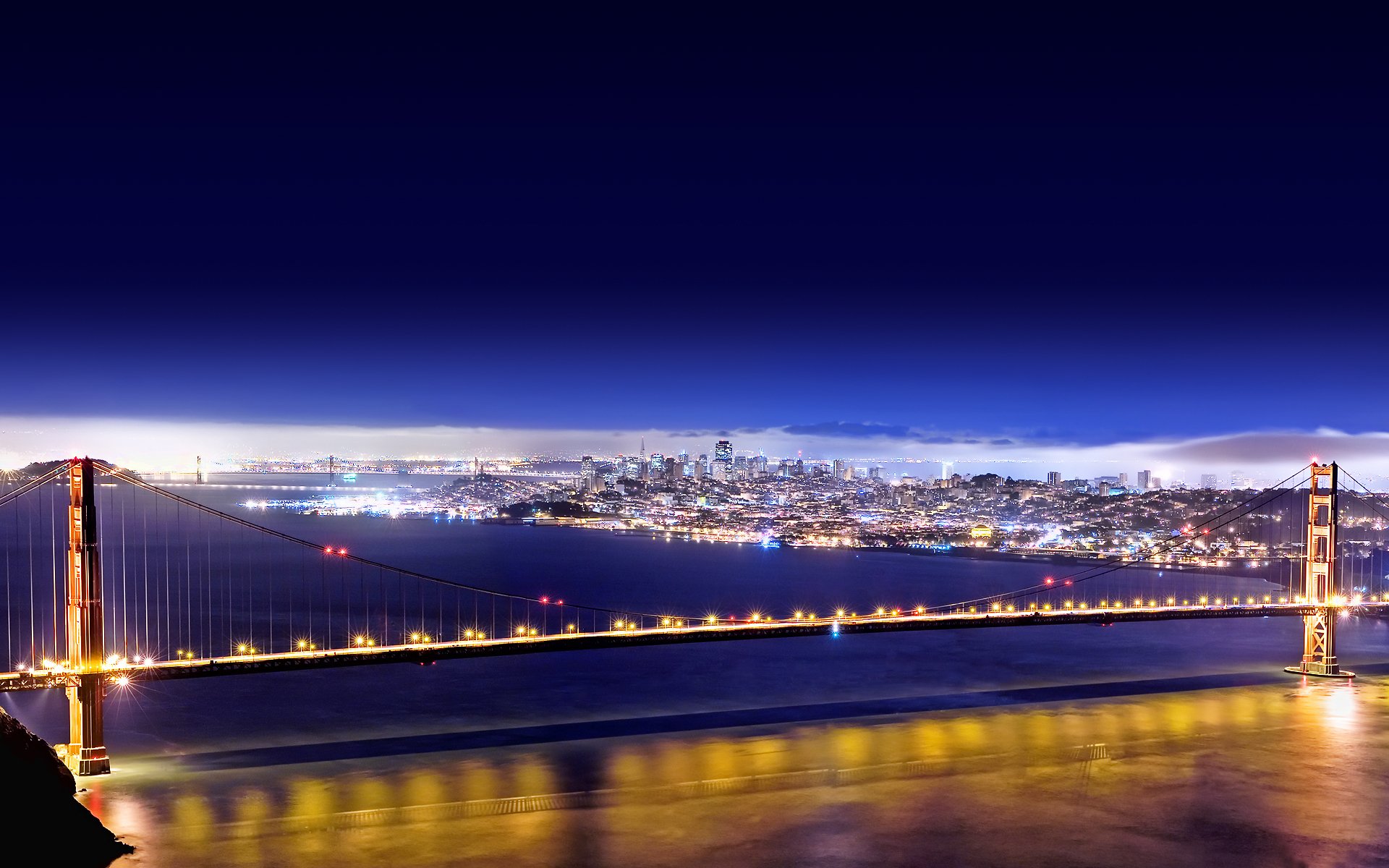 A stunning HD wallpaper showcasing the Golden Gate Bridge at night, illuminated against the backdrop of San Francisco's skyline, reflecting shimmering lights on the water.