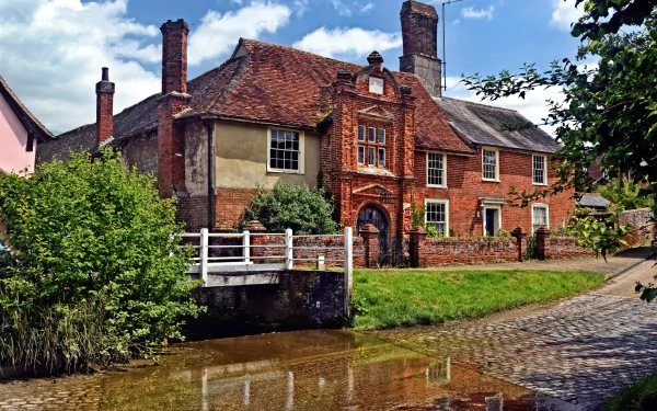 2K Quad HD PC desktop wallpaper and background: man-made historic brick house by a calm canal with a white footbridge, cobblestone path and lush greenery under a blue sky.