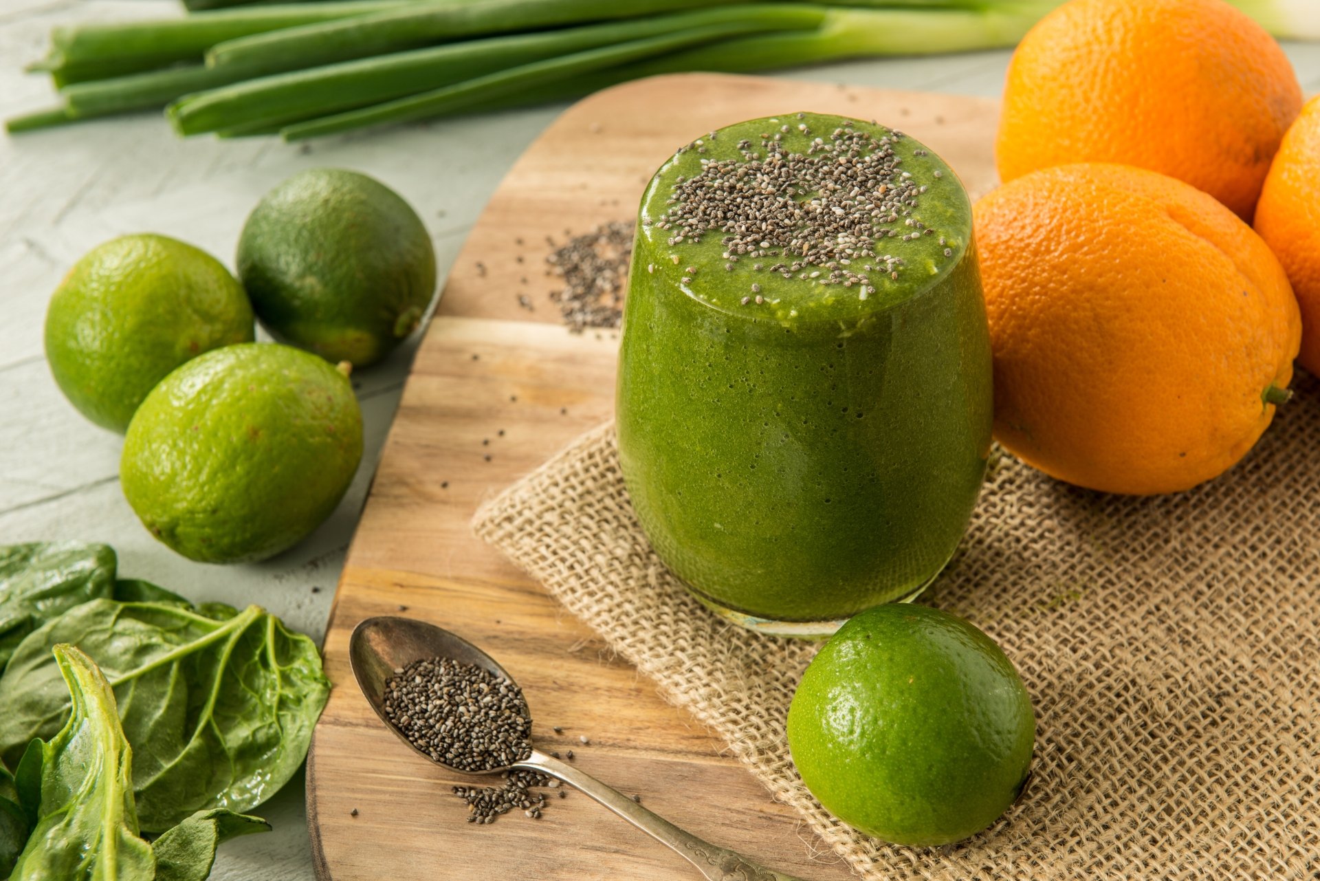 A vibrant green smoothie topped with chia seeds sits on a wooden board surrounded by fresh oranges, limes, spinach, and scattered chia seeds in this 4K Ultra HD desktop wallpaper.