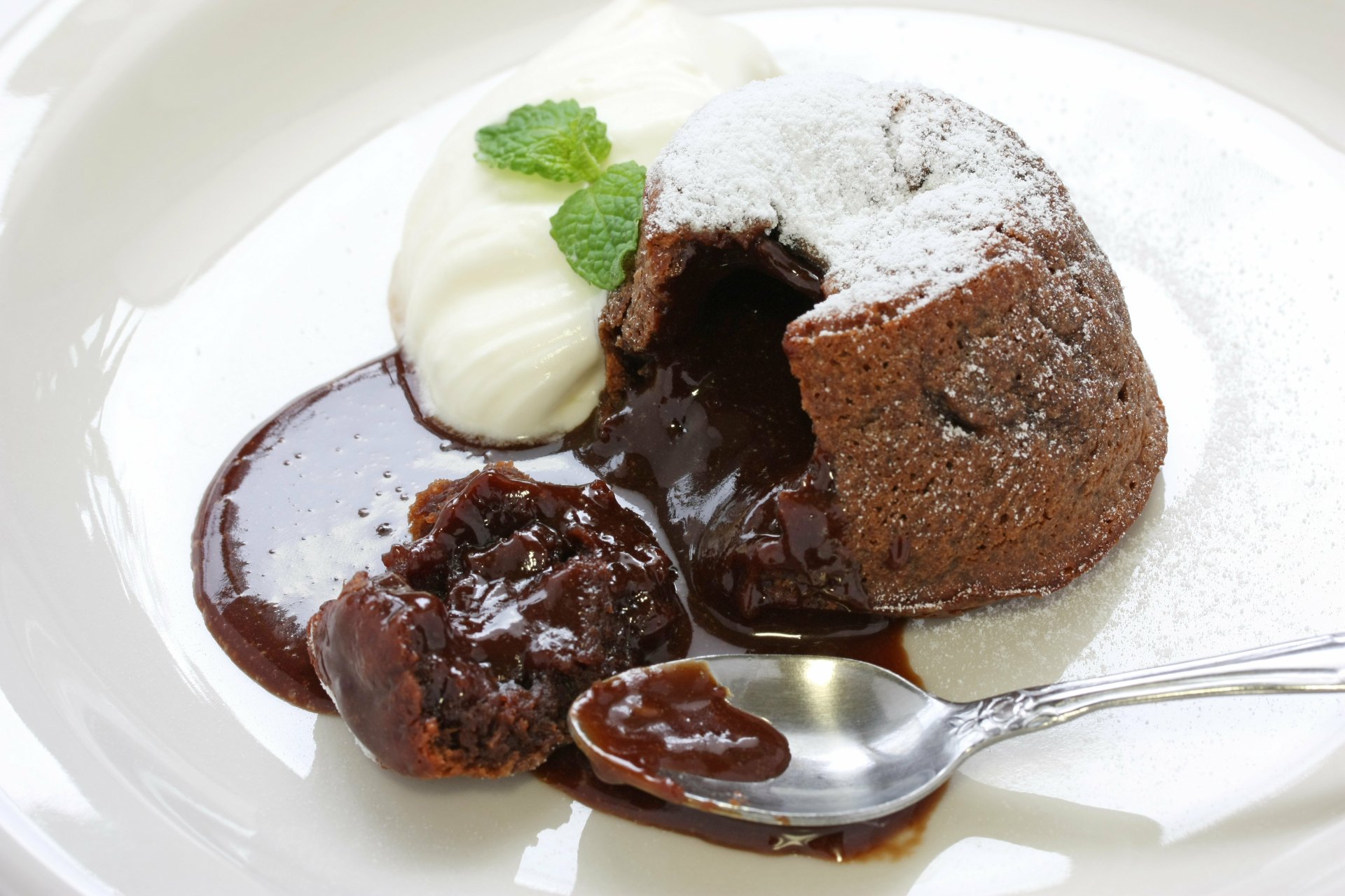 A decadent chocolate lava cake, dusted with powdered sugar, served with a dollop of whipped cream and mint leaves, creating an enticing dessert scene. HD desktop wallpaper.