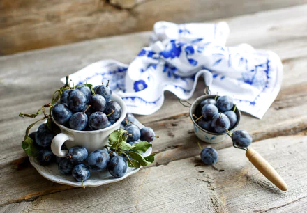 HD PC desktop wallpaper showing ripe plums (fruit, food) heaped in teacups and a small sieve on a rustic wooden table with a blue-and-white cloth background.