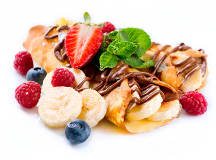 A delightful dessert featuring crêpes topped with bananas, strawberries, mint, and rich chocolate, surrounded by fresh raspberries and blueberries against a bright white background.