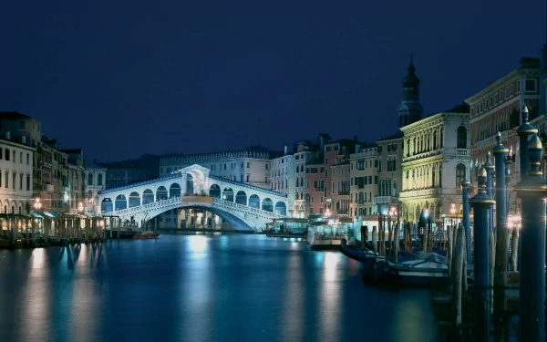 A serene night view of Venice, featuring the illuminated Rialto Bridge over a tranquil canal, surrounded by charming buildings reflecting in the water. A stunning 4K Ultra HD desktop wallpaper.