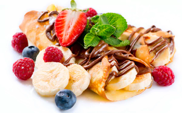 A delightful dessert featuring crêpes topped with bananas, strawberries, mint, and rich chocolate, surrounded by fresh raspberries and blueberries against a bright white background.