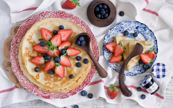 HD desktop wallpaper featuring a delicious crêpe topped with fresh strawberries and blueberries, accompanied by plates and a jar of jam on a white cloth background.