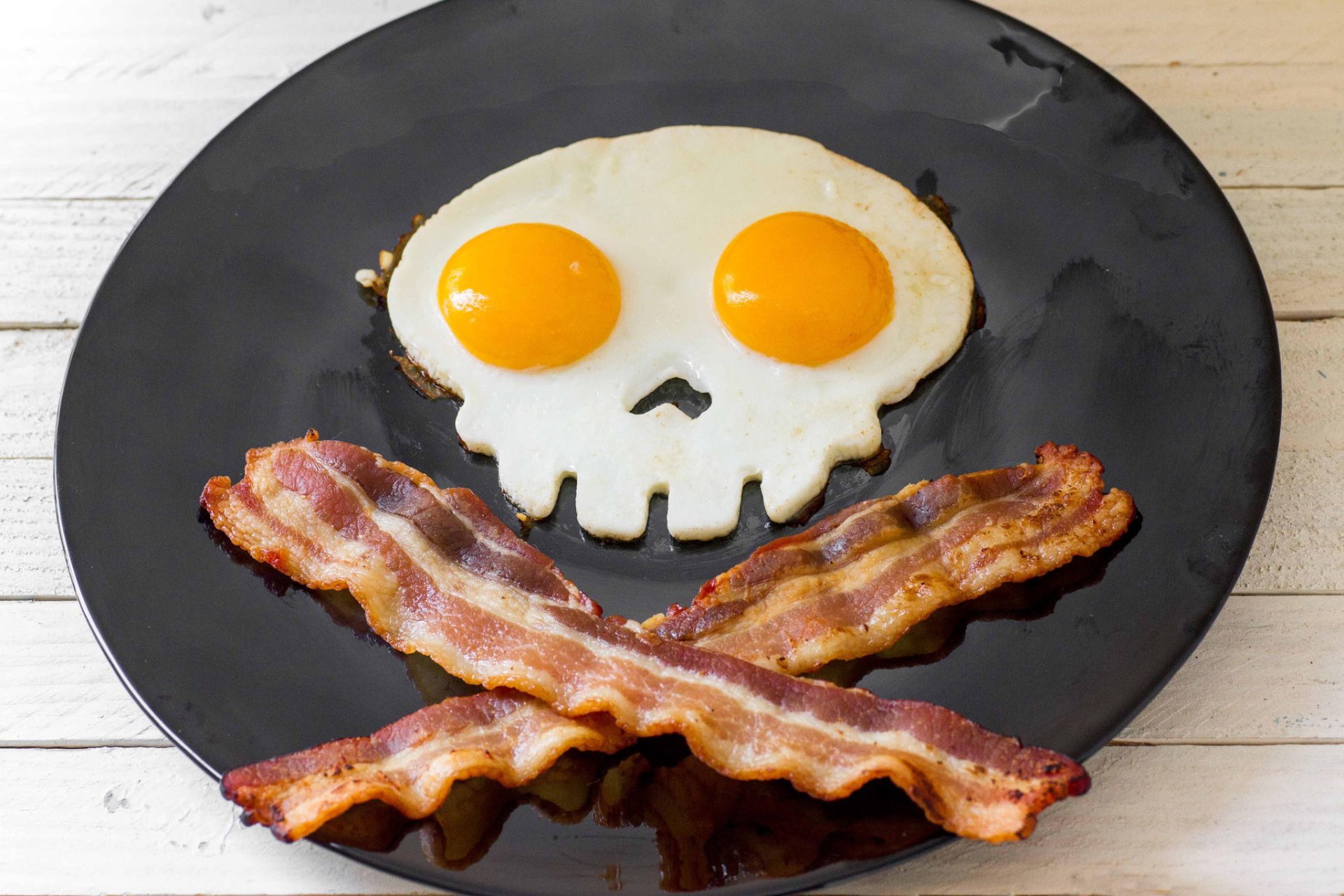 HD desktop wallpaper showing a breakfast plate with two fried eggs shaped like a skull and crossed strips of crispy bacon forming a pirate-style design.