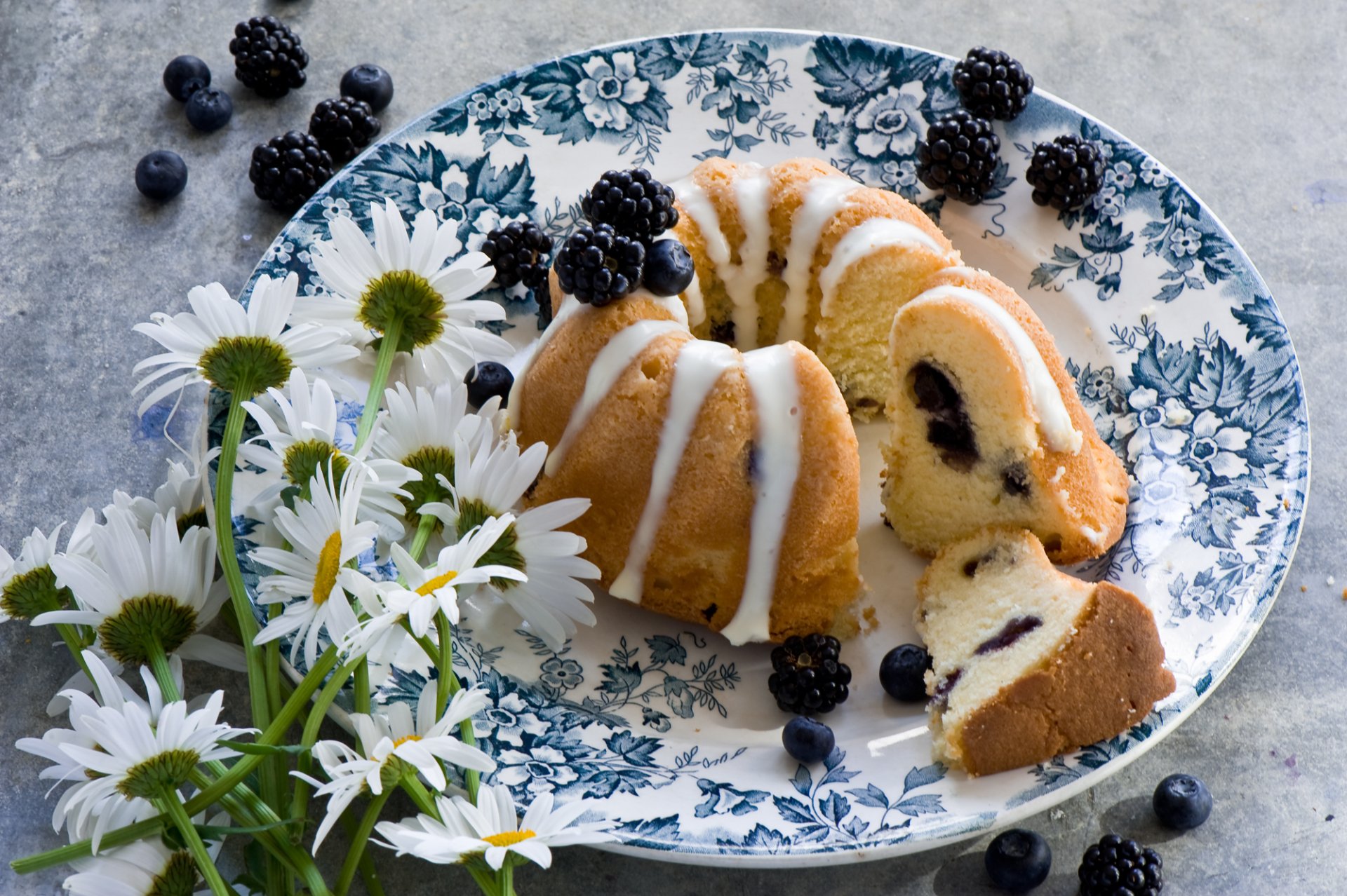 A beautiful floral plate holds a sliced bundt cake with blueberries and blackberries, surrounded by fresh flowers, creating a vibrant and appetizing scene.