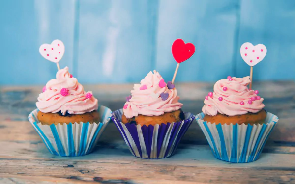 Three pink-frosted muffins with heart-shaped toppers sit on a wooden surface against a blue background, captured in vibrant 4K Ultra HD detail as a cupcake dessert wallpaper.